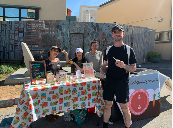 Outdoor market booth with four vendors behind a table of vegan food samples and one person in front holding a cup.